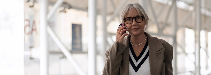 Older woman with glasses talking on the phone while walking in a modern urban setting during the day