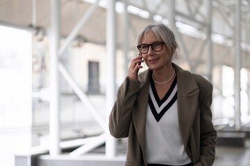 Middle aged businesswoman in stylish coat smiling while talking on the phone in a modern office building during the day