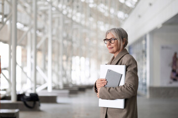 Older woman in a business suit holding a laptop and documents stands confidently in a modern office building during daytime