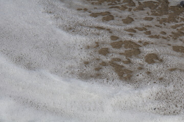 Close-up of foamy sea water washing onto sandy beach, creating textured bubbles and layers of white foam over wet sand
