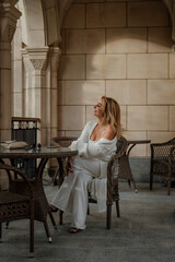 Woman White Outfit Patio Sunlit Stone Wall - A woman in a white outfit sits at a table on a patio with a stone wall in the background, bathed in sunlight.