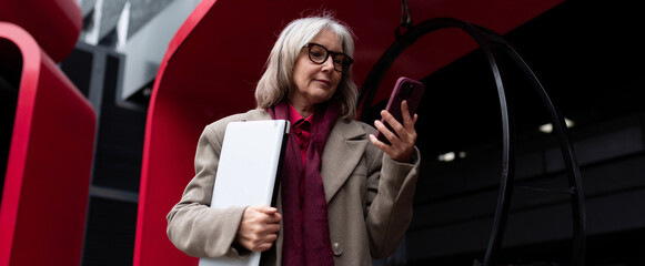 Senior businesswoman checking her smartphone while standing outside in an urban setting during the...
