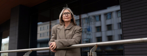 Stylish elderly woman enjoying a sunny day outside a modern building while leaning on a railing in...