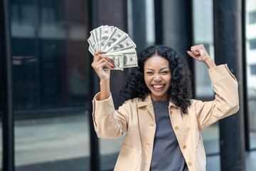 Joyful woman holding a fan of cash in one hand and raising her fist in celebration, expressing financial success and happiness.