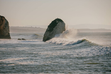 A large rock is in the ocean with a wave crashing over it. The water is calm and the sky is clear