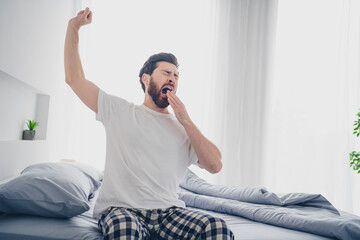 Photo of funny tired young man nightwear pajama sitting bed stretching arms yawning indoors house...