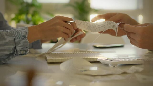Two people are sorting through piles of receipts while using a calculator to manage their finances, surrounded by a bright and welcoming office atmosphere