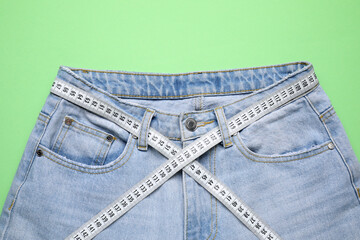 Jeans and measuring tape on green background, top view