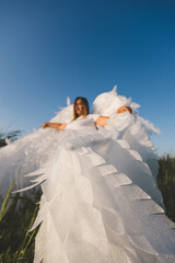 Beautiful angel with huge wings. A woman with large white angel wings stands in a field of green grass under a clear blue summer sky, creating a serene and ethereal scene.