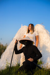 Under a clear blue sky, a masked samurai kneels in the grass holding a katana, and behind him stands an angel with large white wings. The angel protects and guards the warrior samurai.