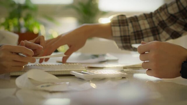 Two individuals are seated at a table surrounded by plants, reviewing receipts and using a calculator to manage their expenses. The atmosphere is light and focused on financial organization