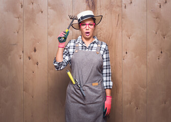 Impressed gardener woman holding rake while looking to camera and say what on wooden background