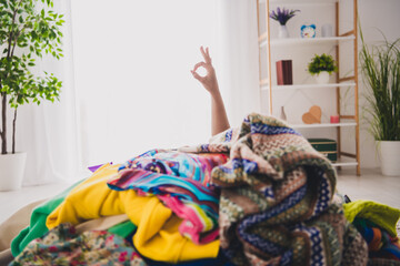 Young woman amidst colorful clothes showing okay sign in bright living room, emphasizing charity...
