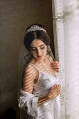 Fototapeta premium A woman in a white dress is standing in front of a window, with her arms crossed and a veil on her head. Concept of elegance and sophistication, as the woman is dressed in a wedding-like outfit