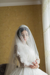 A woman in a wedding dress is standing in front of a yellow wall. She is wearing a veil and a tiara