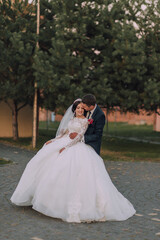 A bride and groom are standing in a park, with the bride wearing a white dress and the groom wearing a suit. They are embracing each other, and the bride is wearing a veil. Scene is happy and romantic