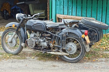 one retro black military iron old with engine and two wheels industrial dirty motorcycle with a sidecar stands on the ground on the street near green fence during the day 	