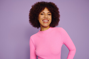 A young African American woman with curly hair radiates joy and confidence in her pink outfit on a purple background.