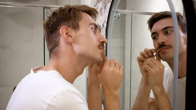 Young man trims his mustache with scissors in the bathroom in front of the mirror.