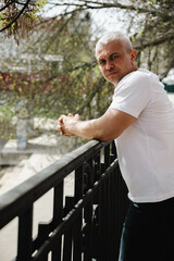 A man in a white shirt stands on a balcony railing. He is looking at the camera with a serious expression