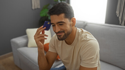 Young man with beard holding credit card sitting on sofa in modern living room with a thoughtful expression