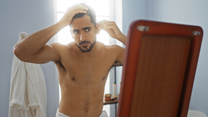 Young hispanic man with a beard examines his reflection in a spa room with calm and focused expression, highlighting grooming and wellness in a serene indoor setting.