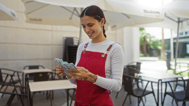 Woman counting money at an outdoor cafe terrace wearing a red apron. - Powered by Adobe