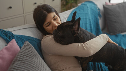 A young hispanic woman cuddles a french bulldog in the living room of her cozy apartment.