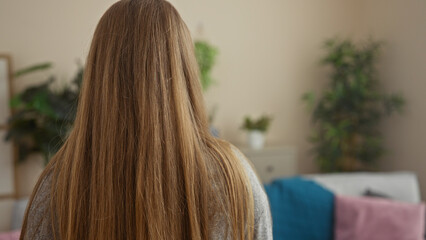 A young blonde woman with long hair is indoors in a living room, facing away, seated in a cozy apartment interior with plants and decorations around her.