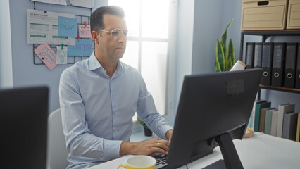 Middle-aged hispanic man working in an office setting on a desktop computer with organized...
