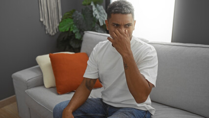 Young man covers nose from bad smell while sitting on sofa in living room at home, displaying discomfort.