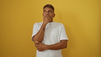 Young hispanic man in deep thought, isolated against a vibrant yellow background, embodying a...