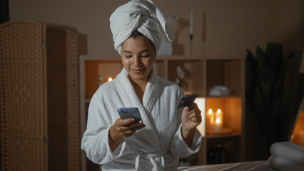 Hispanic woman in a spa wearing a bathrobe and towel, using a mobile phone while holding a credit...