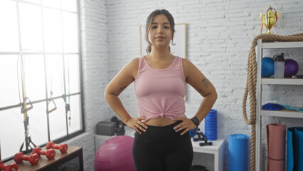 Fototapeta premium Young hispanic brunette woman in a gym wearing a pink top and black leggings, standing confidently with hands on her hips in an indoor sport center with exercise equipment around her.