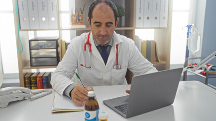 A mature hispanic male doctor in a clinic writes notes while working on a laptop at his workplace desk.