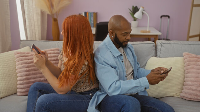 Interracial couple sitting back to back in living room using smartphones, depicting modern relationship dynamics with home comfort