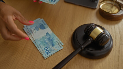 Woman's hand with pink nails counting brazilian reais on a wooden desk with a judge's gavel and a...