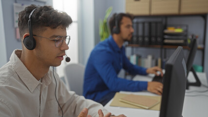 Hispanic men working together in an office, wearing headsets in a business environment, focused on their tasks and collaborating as employees or executives indoors.