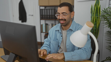 Hispanic man working in an office, typing on a computer and smiling while sitting at a desk with a...