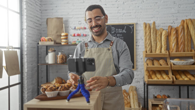 Young man recording video in bakery shop, surrounded by bread and pastries, wearing apron and glasses, smiling, holding smartphone, natural light, cozy indoor setting