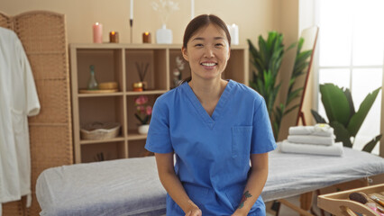 Young chinese woman in blue scrubs smiling at a spa salon with candles and plants in the background