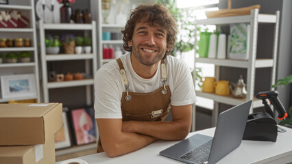 Young man with crossed arms smiling in a decor store, standing behind a counter with a laptop and...