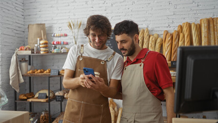 Men smiling in bakery, showing phone in aprons, with bread and pastries on shelves in background,...