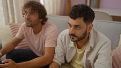 Two men sitting together on a couch in a cozy living room, intently focused on something ahead, suggesting a moment of bonding and shared activity.