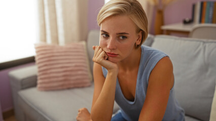 Young woman with short blonde hair sitting in a living room and looking thoughtful in a cozy home...