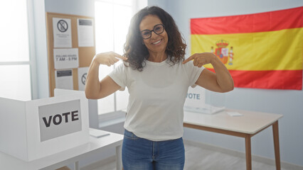 Woman voting in spanish election smiling and pointing at herself in a voting room with spanish flag in the background