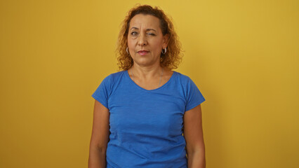 Mature hispanic woman with curly hair winking while standing over isolated yellow background.