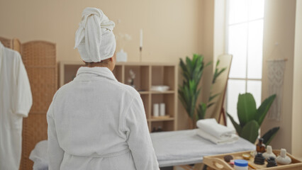 A middle-aged hispanic woman in a white robe and towel in a serene spa room with calming decor, soft lighting, plants, and wellness products.