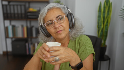Woman listening with headphones in an office wearing glasses and holding a cup, surrounded by bookshelves, plants, and office decor, conveying a focused and thoughtful mood