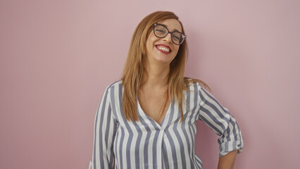 Fototapeta premium Woman smiling confidently, standing against a pink wall, middle aged with blonde hair and glasses, isolated pink background, striped blouse, mature female, cheerful expression